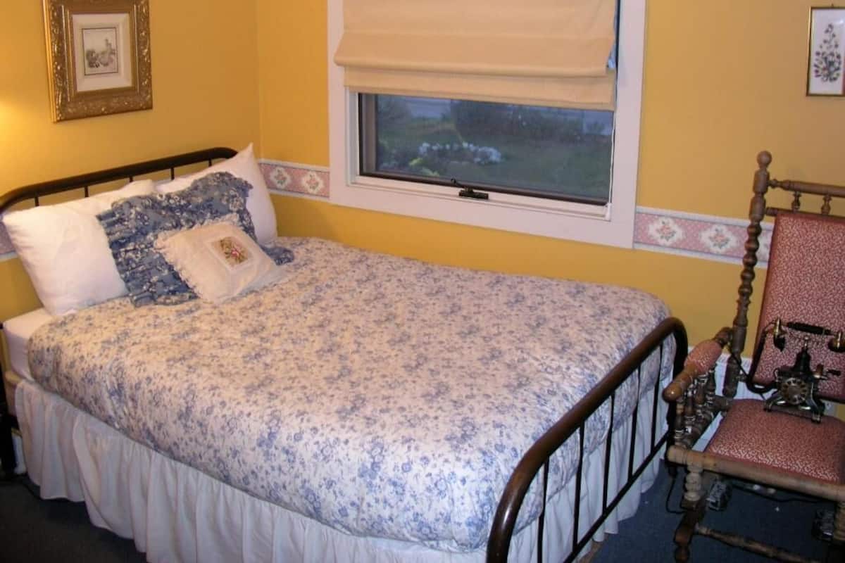 A single bed with a floral bedspread, a window with a blind, a wooden chair, and a picture frame on the wall.
