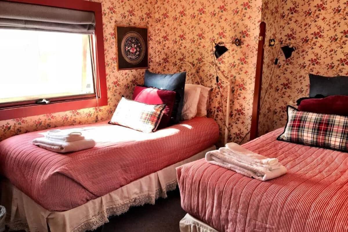 A room with two beds, floral wallpaper, a window, and a framed picture on the wall.
