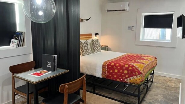 Ein kleines Zimmer mit einem Bett, einem Schreibtisch mit Stuhl, einer Pendelleuchte und einer Klimaanlage.