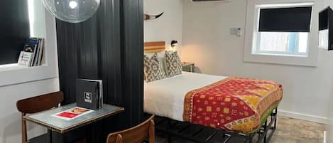 Quarto pequeno com cama, escrivaninha com cadeira, luminária pendente e ar condicionado.