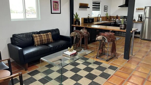Un salon avec un canapé en cuir noir, une table basse en verre et un tapis à carreaux. On aperçoit une cuisine à l’arrière-plan, équipée d’appareils électroménagers modernes.