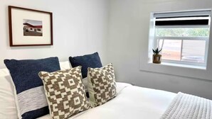 Une chambre à coucher avec un lit, deux oreillers, un tableau encadré au mur, une fenêtre avec des stores et une petite plante sur le rebord de la fenêtre.