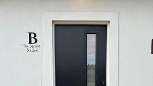 Une porte noire avec un panneau de verre, un mur blanc orné des lettres « B » et de l’inscription « The Service Station », et un baril rouillé portant les étiquettes « TEXACO » et « MOLYXIE CHEESE ».