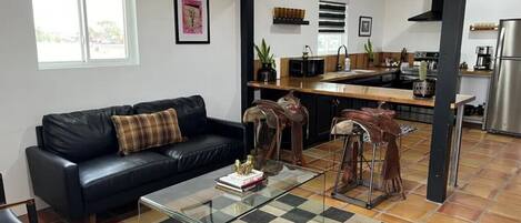 A living room with a black leather sofa, a glass coffee table, and a checkered rug. There is a kitchen visible in the background with modern appliances.