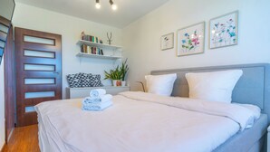 A neatly made bed with white linens, a grey headboard, and a bedside table with a plant. There are bookshelves above the bed and three framed floral artworks on the wall. - Urban Chic Apartment (Katowice)
