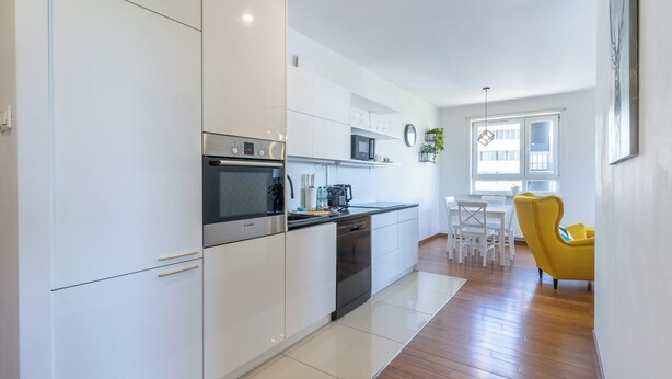 A modern kitchen with white cabinets, a Bosch oven, and a microwave. There is a dining area with a yellow chair and a white table. - Urban Chic Apartment (Katowice)