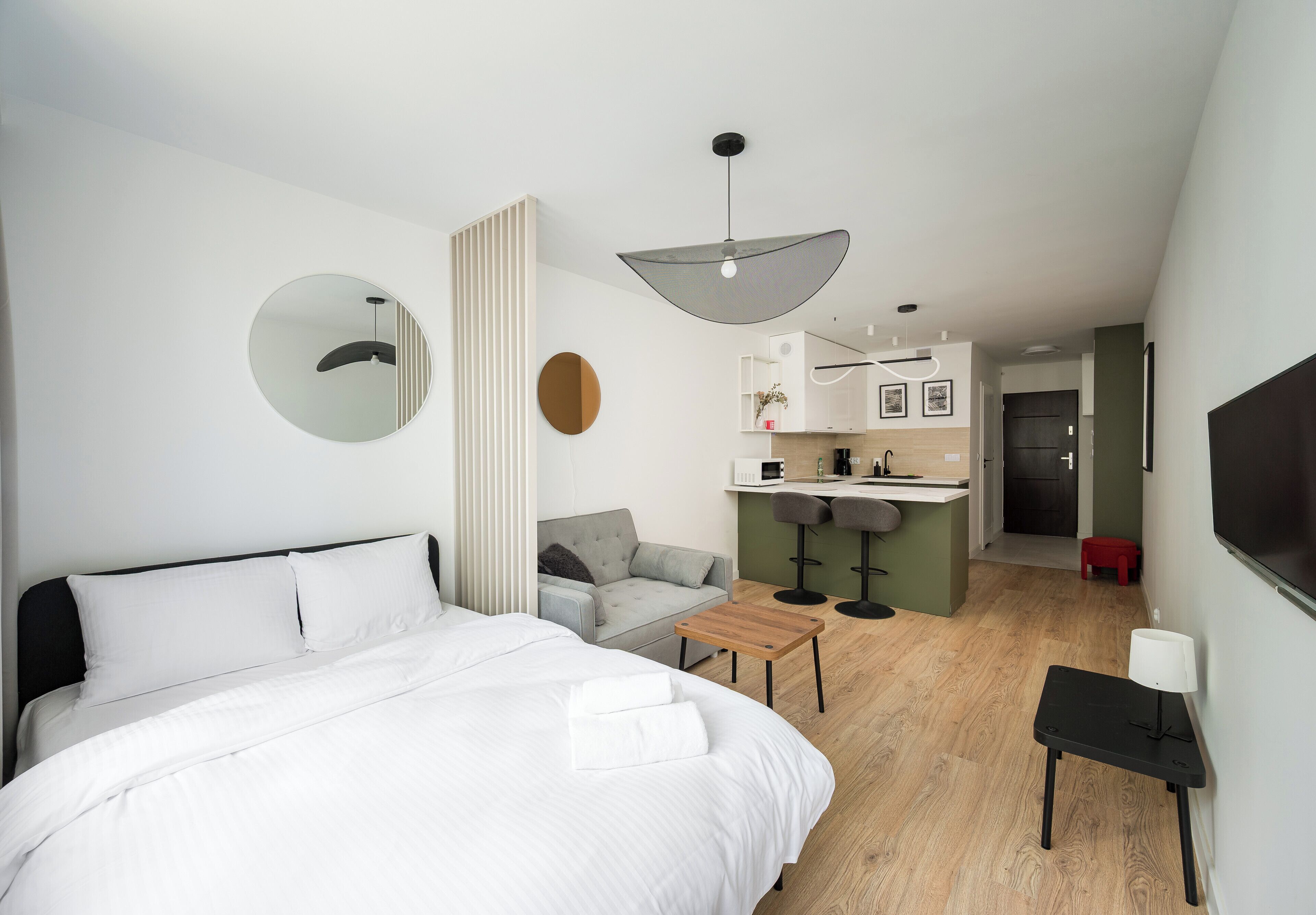 A modern hotel room with a large bed, a small desk, a sofa, a kitchen area, and a TV.