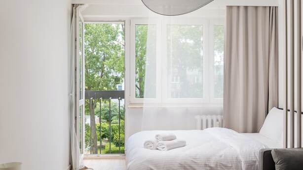 Een slaapkamer met een bed, een stoel, een raam met gordijnen en uitzicht op groen.