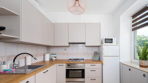 A modern kitchen with white cabinets, a wooden countertop, a gas stove, an oven, a microwave, and a window with blinds. - Metro Młynów Comfort Apartment (Warszawa)