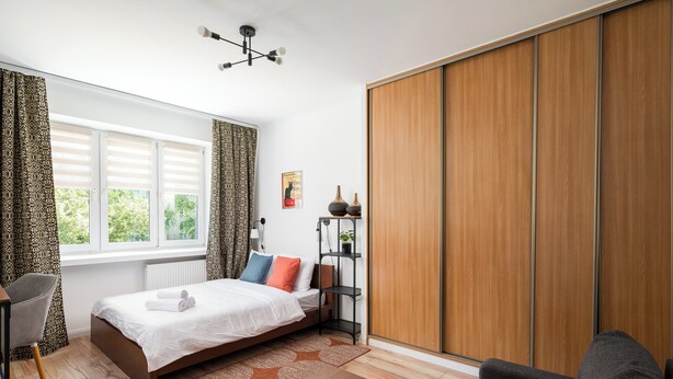 A modern bedroom with a bed, a desk, a chair, a wardrobe, and a window with curtains. - Metro Młynów Comfort Apartment (Warszawa)