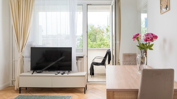 A modern living room with a flat-screen TV, a dining table with chairs, a vase with flowers, and a balcony with a chair. - Złota Warsaw Skyline Apartment (Warszawa)
