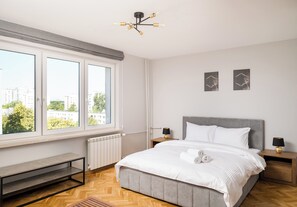 A modern bedroom with a large window, a bed with white bedding, two bedside tables with lamps, and a bench by the window. - Wola Smocza Comfort Studio (Warszawa)