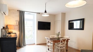 Un coin repas avec une table en bois et des chaises blanches, un téléviseur fixé au mur et une fenêtre avec des rideaux.