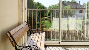 Een balkon met een houten bankje, uitzicht op een tuin, en een gebouw op de achtergrond.