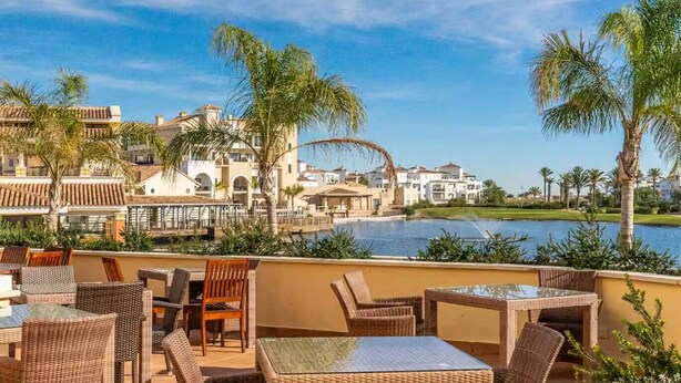 A resort with outdoor seating, a pool, and buildings in the background.