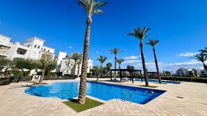 A swimming pool with palm trees, a pergola, and a building with balconies in the background.