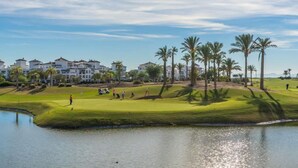 A golf course with a water body, palm trees, and buildings in the background.