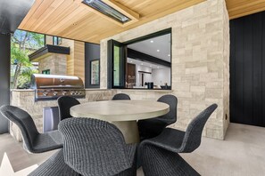 An outdoor dining area with a round table and wicker chairs, a built-in grill, and a stone wall.