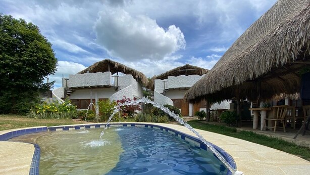 Una piscina con fuente, rodeada de edificios con techo de paja y un cielo despejado.