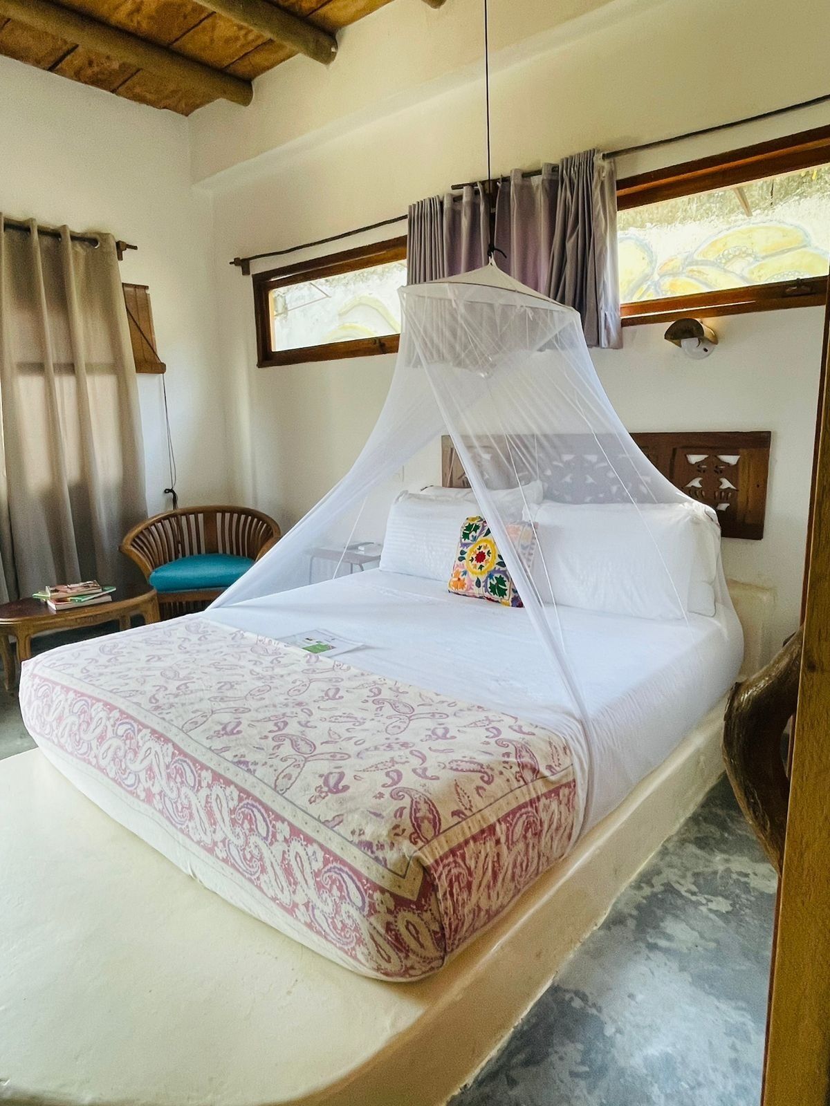 Una cama individual con dosel, una mesita de noche y una silla en una habitación con techo de madera y ventana con cortinas.