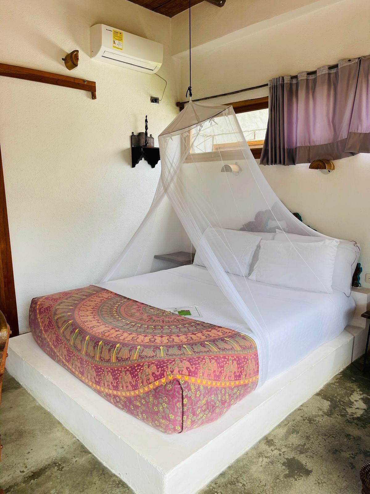 Una habitación con una cama bajo una mosquitera, un aire acondicionado blanco y una ventana con cortinas.