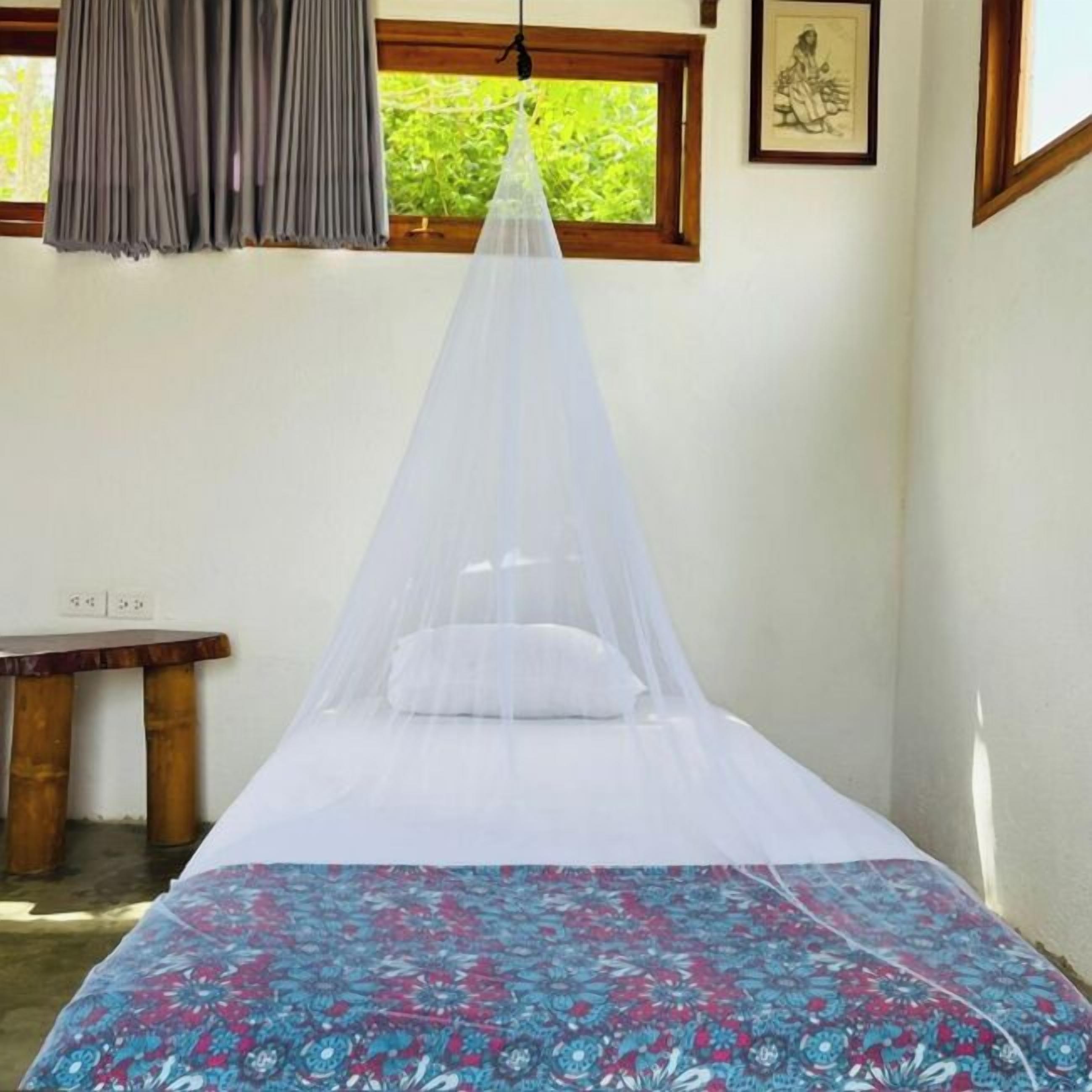 Une chambre avec un lit recouvert d’un dais blanc, une couvre-lit à motifs, un banc en bois et une fenêtre donnant sur un paysage verdoyant.