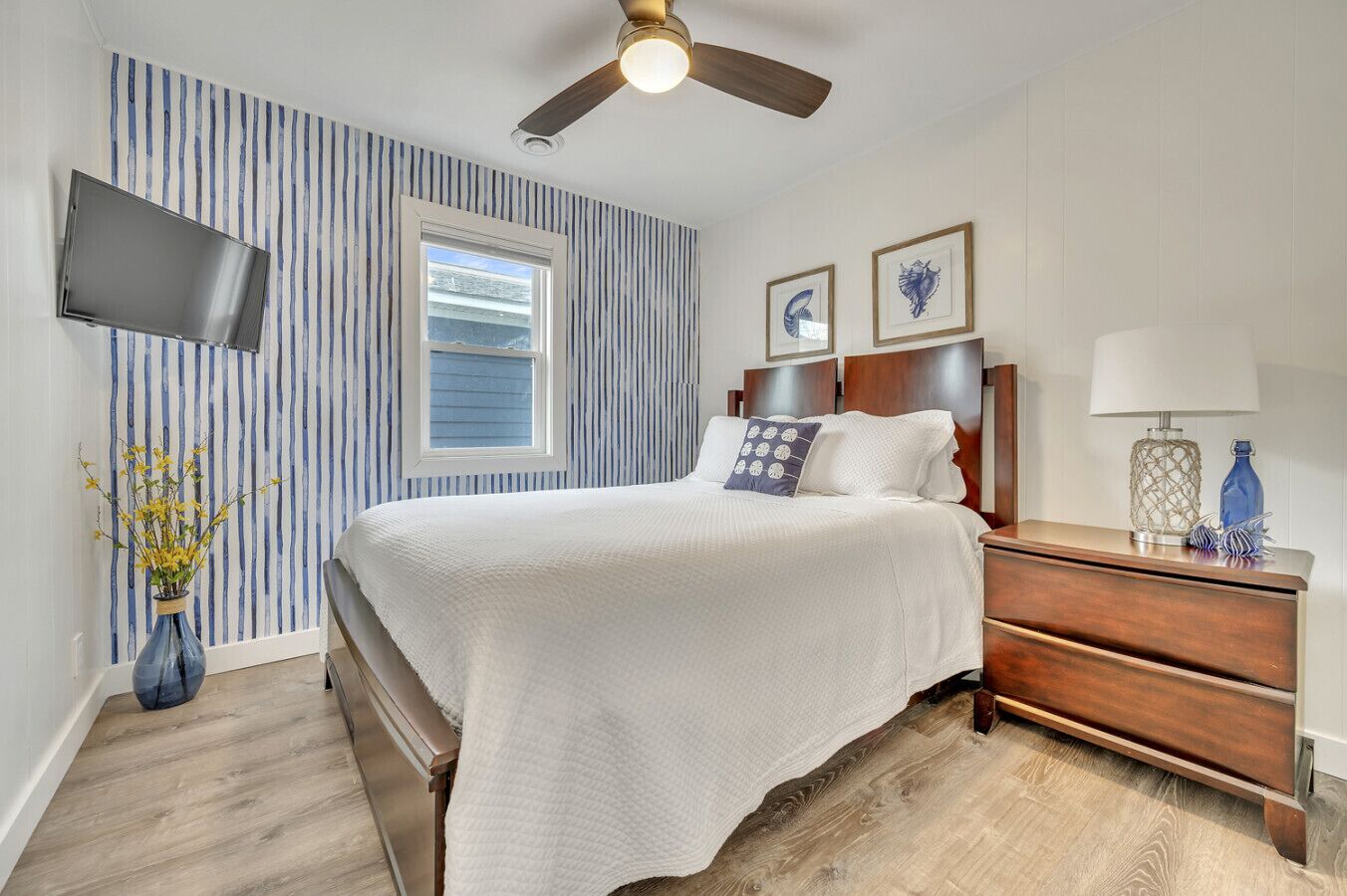 A bedroom with a bed, a nightstand, a lamp, a vase with flowers, a ceiling fan, a TV, and a window.