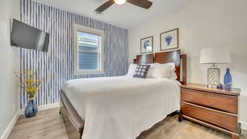 A bedroom with a bed, a nightstand, a lamp, a vase with flowers, a ceiling fan, a TV, and a window.