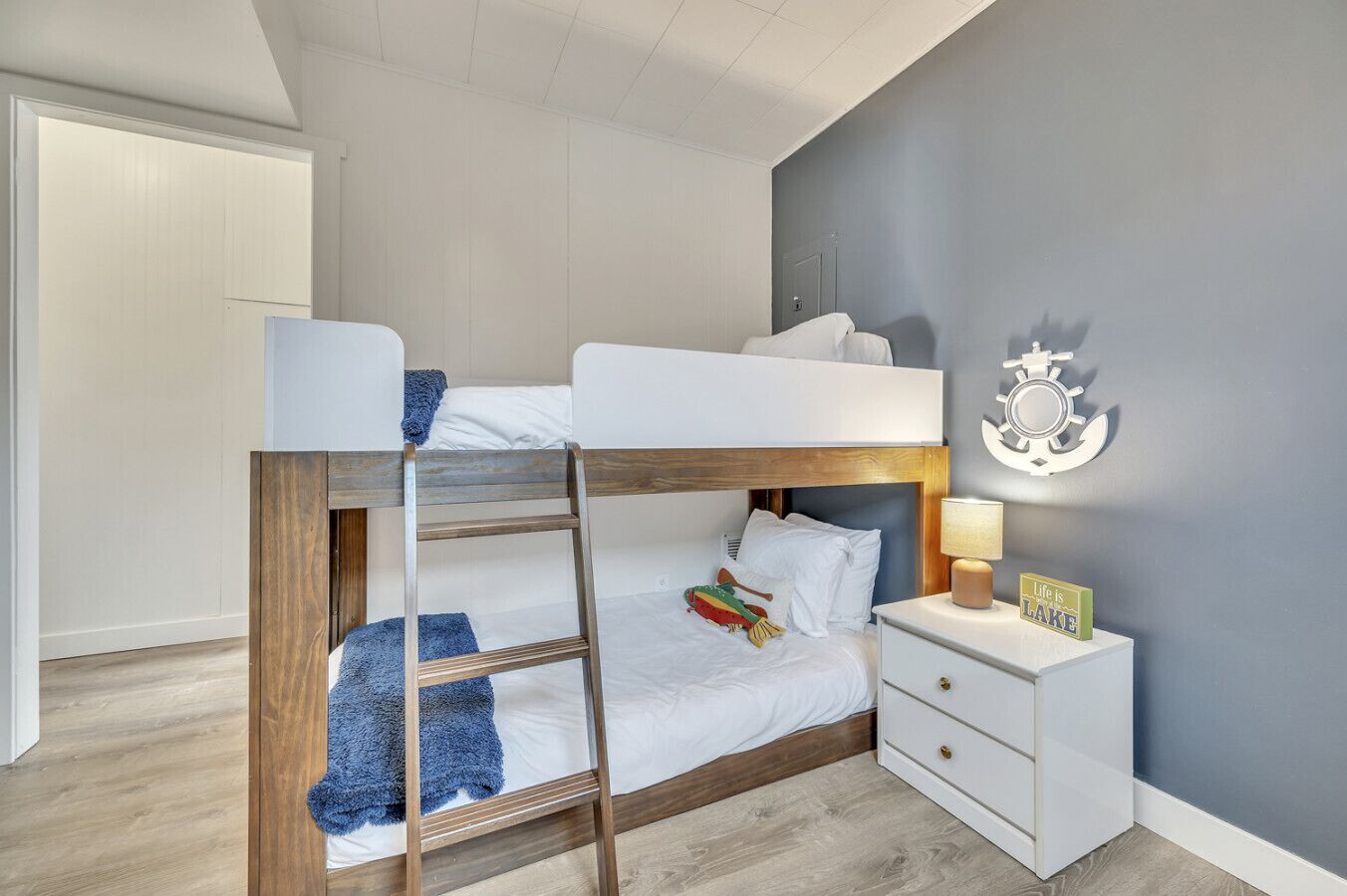 A bunk bed with a wooden frame, white bedding, and a blue throw blanket. There is a bedside table with a lamp and a decorative item. The room has light-colored walls and a wooden floor.