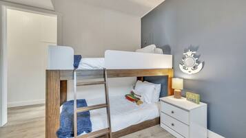 A bunk bed with a wooden frame, white bedding, and a blue throw blanket. There is a bedside table with a lamp and a decorative item. The room has light-colored walls and a wooden floor.