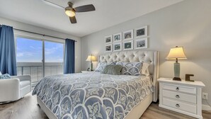 A bedroom with a large bed, a ceiling fan, a nightstand with a lamp, a chair, and a window with curtains. - NEW LISTING...AWave From It All - Lakefront on Big Spirit Lake (Orleans)