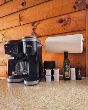 Una cafetera Keurig sobre una encimera con un dispensador de toallas de papel, una botella de aceite de oliva y varios recipientes pequeños.