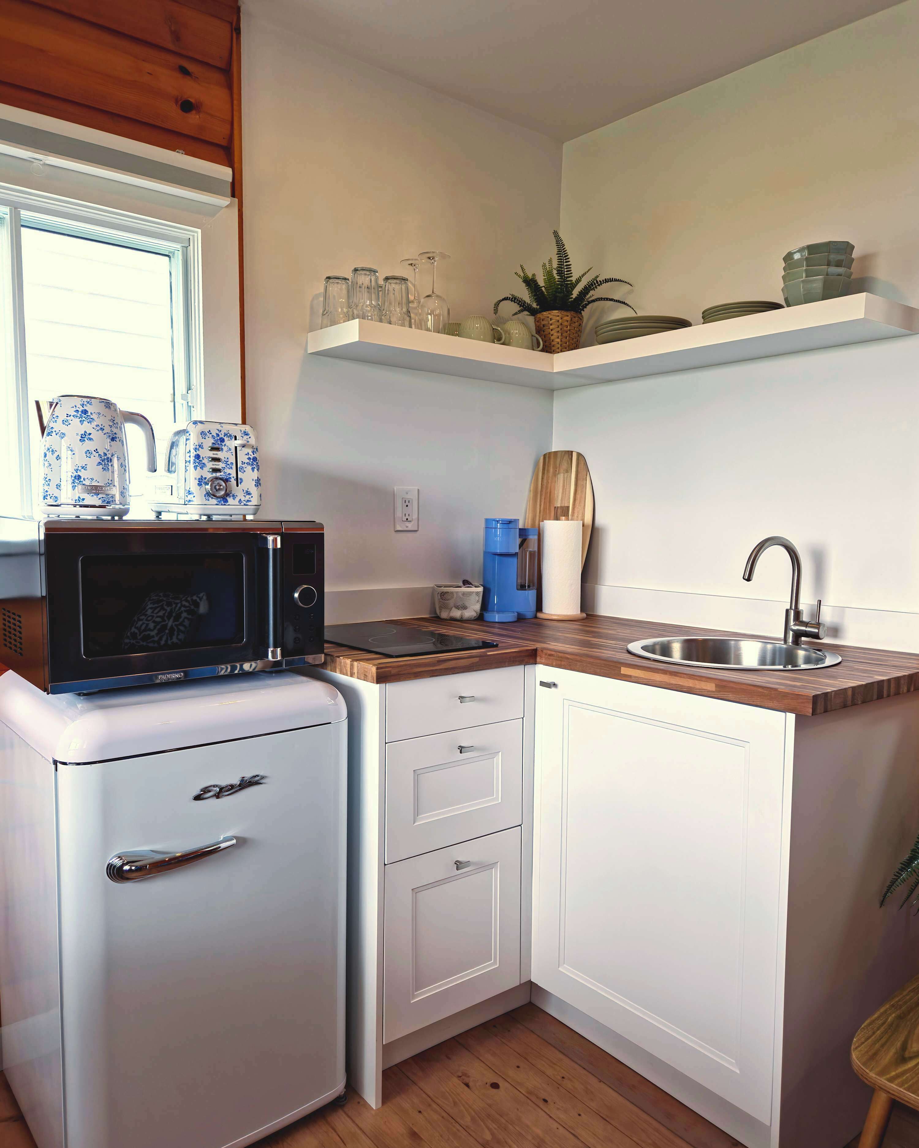 Et lille køkken med mikroovn, toaster og minikøleskab. Der er en vask med moderne armatur, et vindue og en hylde med service og en plante.