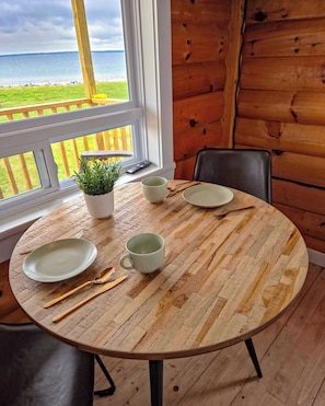 A round wooden table with plates, cutlery, and a cup, set against a wooden wall with a window overlooking a grassy area. - Cottage # 7 Hunts Point Beach Cottages 1 Bedroom (Hunts Point)