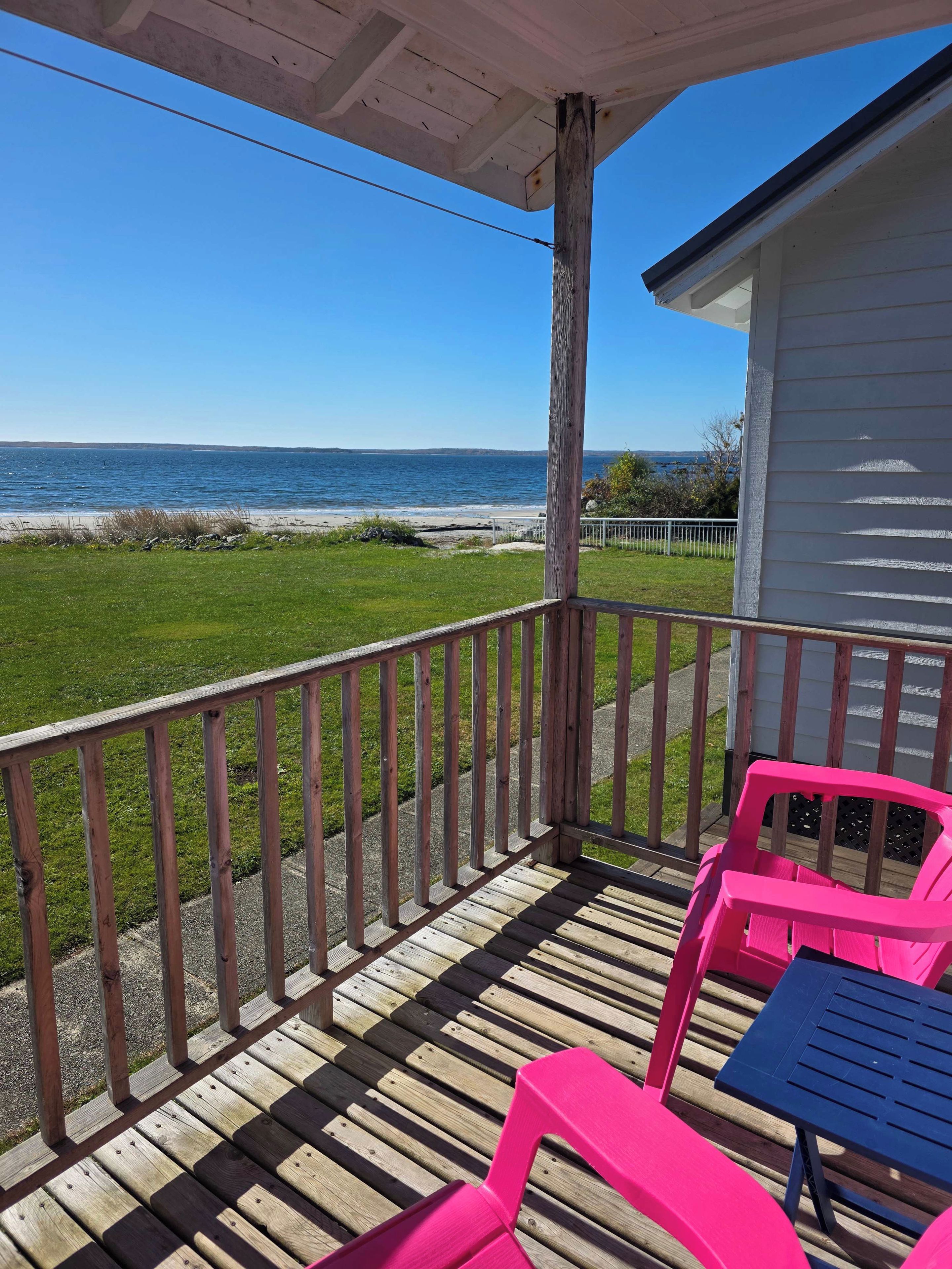 A wooden deck with a railing, a table, and two chairs, overlooking a grassy area and a body of water.