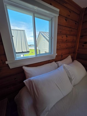 A wooden cabin room with a bed, pillows, and a window overlooking a house and a grassy area.
