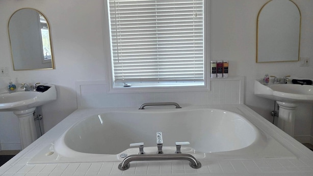 Un baño con una bañera grande, dos lavamanos, una ventana con persianas y un espejo.