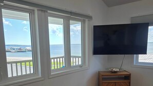 A living room with a large TV, two windows with a view of the water, a wooden cabinet, and a leather sofa. - Pent House at Hunts Point Beach Cottages (Hunts Point)