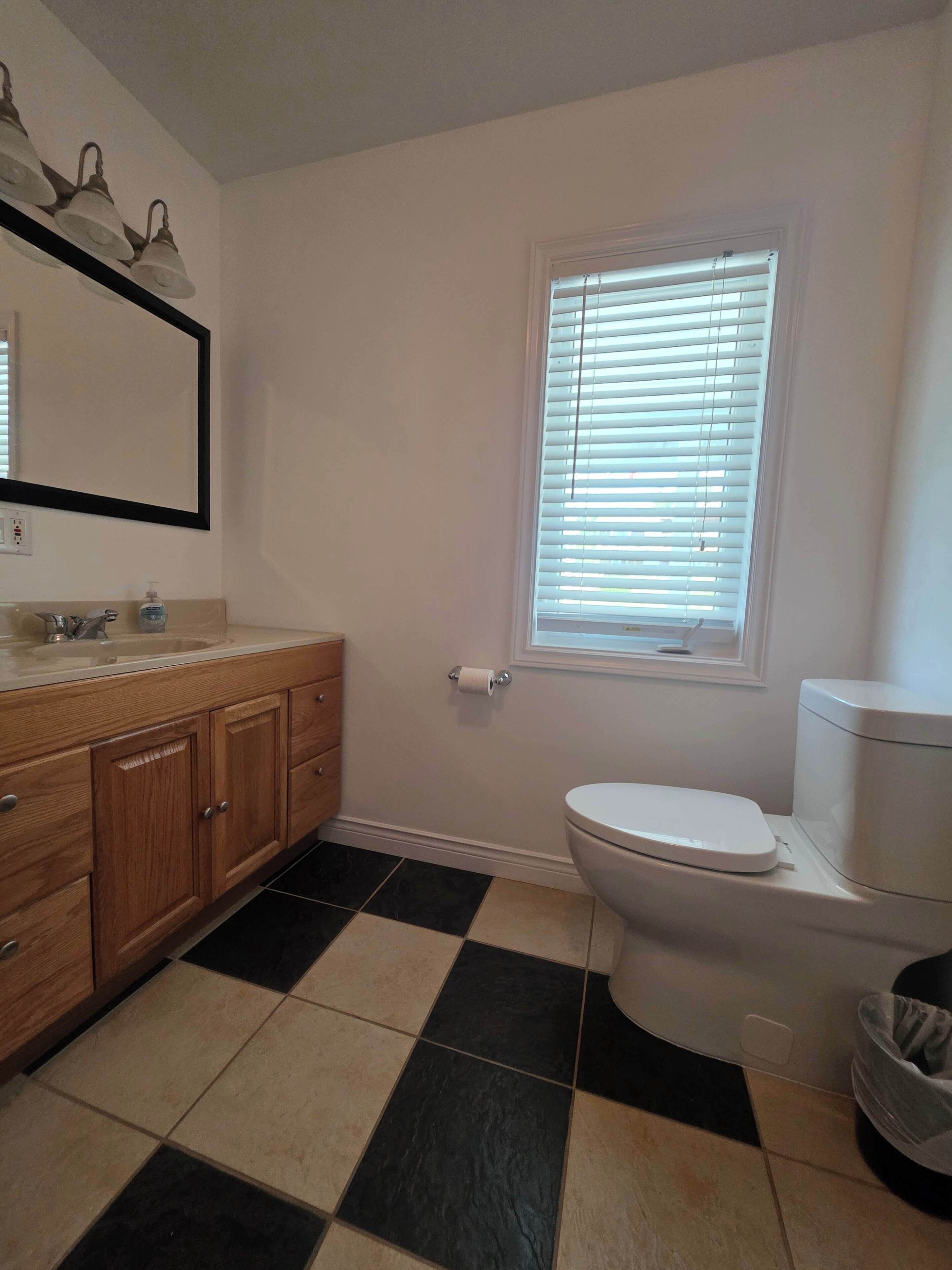 Une salle de bain comprenant des toilettes, un lavabo, un miroir et une fenêtre avec des stores.
