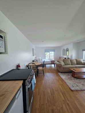 A modern living room with a sofa, a dining table with chairs, a kitchen area with a stove, and a large window providing natural light.