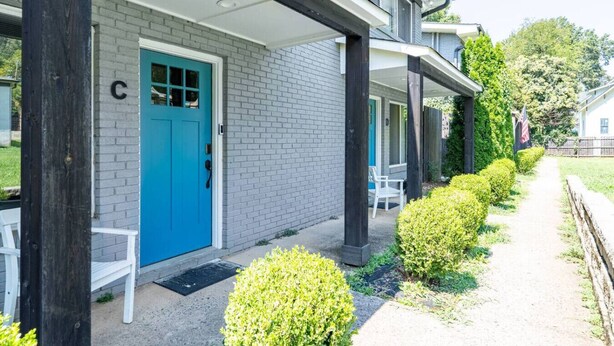 Een gebouw met een blauwe deur, een overdekte veranda en een pad dat naar een witte stoel leidt.