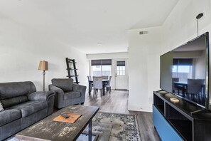 Uma sala de estar moderna com sofá, mesa de centro, televisão e uma área de refeições ao fundo.