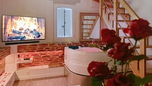A room with a brick wall, a large TV displaying a fire, a bathtub, and a wooden staircase.