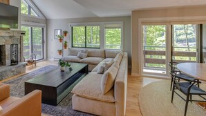 A spacious living room with a large sofa, a coffee table, and a fireplace. There are large windows providing natural light and views of the outdoors.