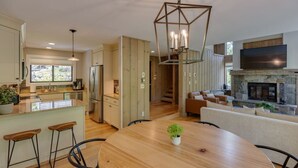 A modern kitchen with a central island, bar stools, and a dining table. There is a fireplace and a TV mounted on the wall.