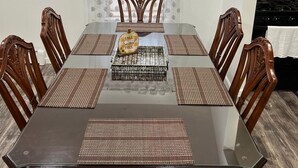 Un comedor con una mesa de vidrio, seis sillas de madera, un centro de mesa decorativo y un ventanal con cortinas de gasa.