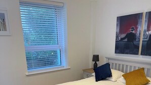 A neatly made bed with white linens and two pillows, a bedside table with a lamp, a window with blinds, and a painting on the wall. - Modern Split-Level 2-Bed Apartment with Balcony (Causeway Coast and Glens)