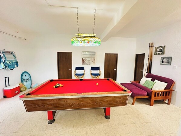 Una mesa de billar con bolas, un sofá rojo, dos sillas plegables y una lámpara colgante en una habitación con paredes blancas y puertas de madera.