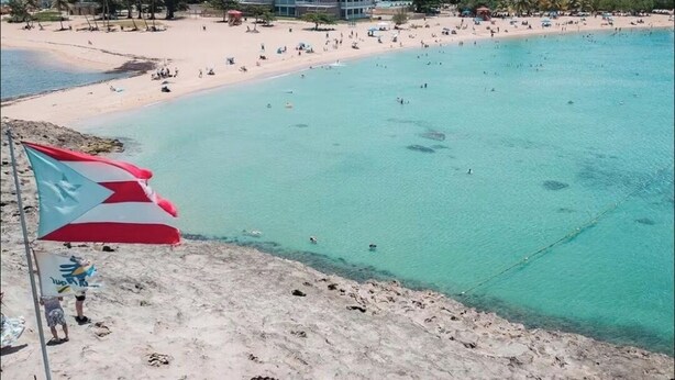 A beach with clear turquoise water, a flag, and a building in the background.