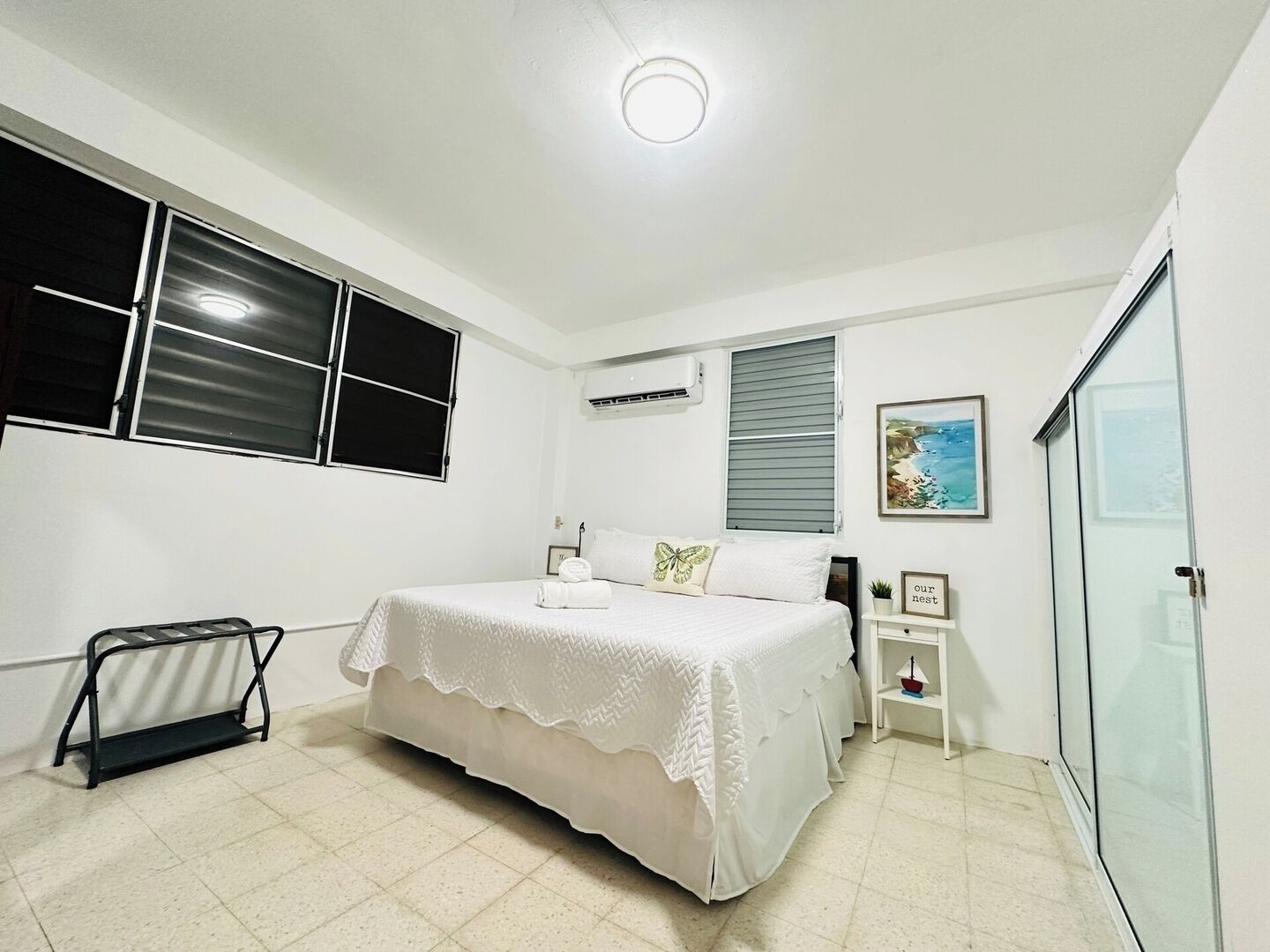 A bedroom with a bed, a bedside table, a wall-mounted air conditioner, a window with blinds, and a framed picture on the wall.